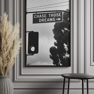 Może przedstawiać: Czarno-biała oprawiona grafika przedstawiająca znak drogowy z napisem "CHASE THOSE DREAMS" i strzałką. Obraz zawiera sygnalizację świetlną, drzewo i pochmurne niebo. Grafika jest wyświetlana na szarej ścianie.