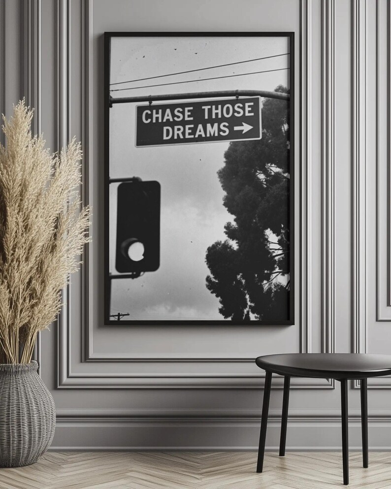 Może przedstawiać: Czarno-biała oprawiona grafika przedstawiająca znak drogowy z napisem "CHASE THOSE DREAMS" i strzałką. Obraz zawiera sygnalizację świetlną, drzewo i pochmurne niebo. Grafika jest wyświetlana na szarej ścianie.