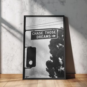 Może przedstawiać: Czarno-biała oprawiona grafika z napisem "CHASE THOSE DREAMS" ze strzałką. Obraz zawiera sygnalizację świetlną i drzewo na tle pochmurnego nieba. Rama jest czarna, a grafika jest wyświetlana na drewnianej podłodze.