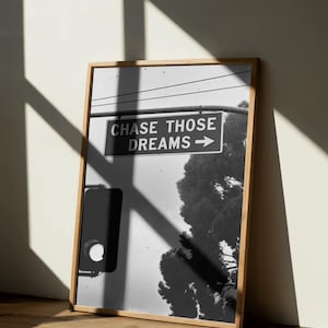 Może przedstawiać: Czarno-biała oprawiona grafika z szyldem ulicznym z napisem "CHASE THOSE DREAMS" ze strzałką w prawo. Obraz zawiera drzewo i światła drogowe. Rama wykonana jest z jasnego drewna.