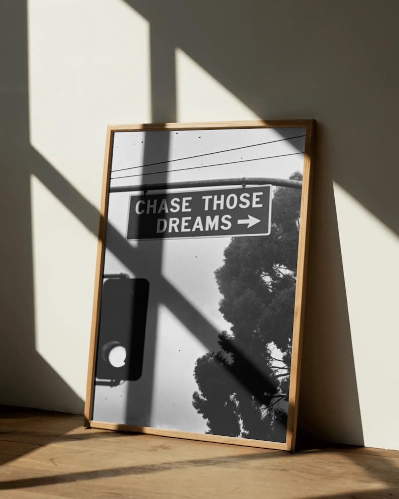 Może przedstawiać: Czarno-biała oprawiona grafika z szyldem ulicznym z napisem "CHASE THOSE DREAMS" ze strzałką w prawo. Obraz zawiera drzewo i światła drogowe. Rama wykonana jest z jasnego drewna.