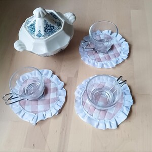 Op de afbeelding: Een witte keramische serveerschaal met een blauw bloemmotief staat naast drie doorzichtige glazen bekers met metalen handvatten, elk op een roze en witte geruite onderzetter met een witte gerimpelde rand. De items zijn gerangschikt op een lichtbruin oppervlak.