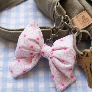 May include: A pink and white patterned bow-shaped keychain with a silver keyring and several keys. The bow is attached to a silver clasp, and the background includes a light-colored bag and a blue and white checkered pattern.
