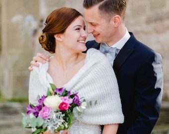 May include: A bride and groom are holding hands and looking at each other. The bride is wearing a white wedding dress with a white knitted shawl and a bouquet of purple and pink flowers. The groom is wearing a dark suit and a white bow tie.