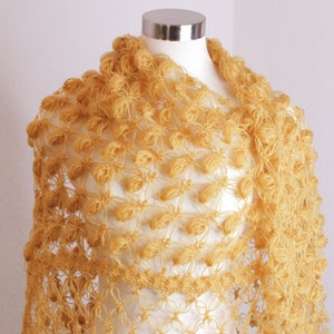 May include: A handmade, mustard-yellow shawl with a unique openwork design. The shawl is draped over a mannequin, showcasing its intricate floral-like pattern and textured, knotted details. The garment is made of yarn.
