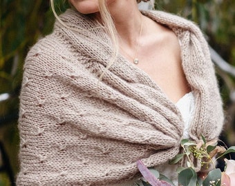 May include: A bride wearing a white lace wedding dress with a brown knitted shawl and gloves. She is holding a bouquet of flowers with pink, white, and green accents.