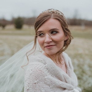 May include: A bride wearing a white knitted shawl and a veil, with a gold and silver tiara in her hair. She is looking off to the side and smiling.