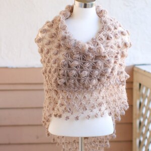 May include: A beige, handmade shawl with a unique, textured design. The shawl is displayed on a mannequin, showcasing its intricate, open-weave pattern and decorative bobbles. The garment's color is a soft, neutral tone, suitable for various outfits.