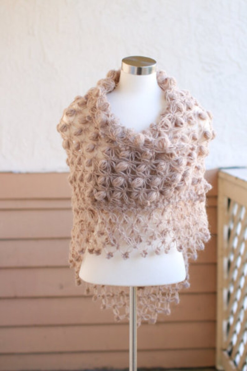May include: A beige, handmade shawl with a unique, textured design. The shawl is displayed on a mannequin, showcasing its intricate, open-weave pattern and decorative bobbles. The garment's color is a soft, neutral tone, suitable for various outfits.