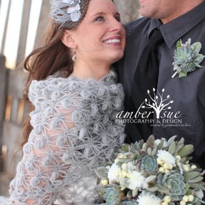 May include: A bride and groom pose for a wedding photo. The bride wears a gray, crocheted shawl and a headpiece with a veil. She holds a bouquet of white and green flowers. The groom wears a gray suit with a succulent boutonniere. The image includes the text "amber sue PHOTOGRAPHY & DESIGN."