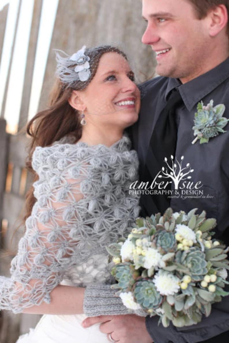 May include: A bride and groom pose for a wedding photo. The bride wears a gray, crocheted shawl and a headpiece with a veil. She holds a bouquet of white and green flowers. The groom wears a gray suit with a succulent boutonniere. The image includes the text "amber sue PHOTOGRAPHY & DESIGN."