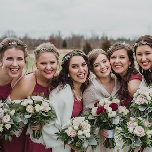 May include: Six bridesmaids wearing burgundy dresses and white shawls, smiling and holding bouquets of white and burgundy roses with greenery.