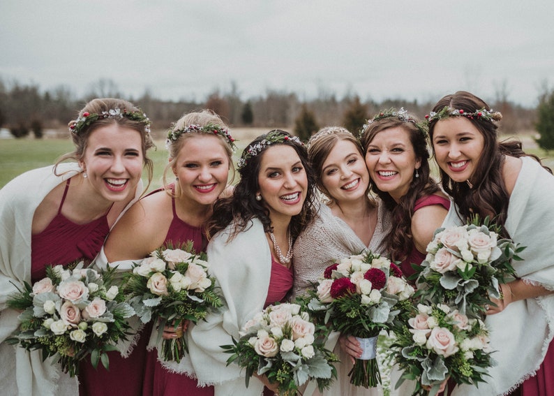 May include: Six bridesmaids wearing burgundy dresses and white shawls, smiling and holding bouquets of white and burgundy roses with greenery.