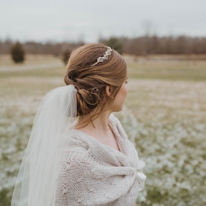 May include: A bride wearing a white knitted shawl and a veil, with her hair styled in a bun and a gold headband. She is standing in a field with a blurred background.