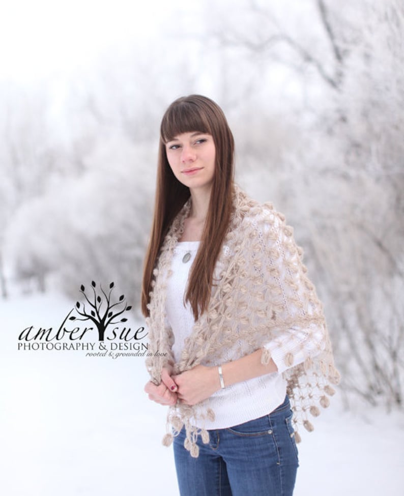 May include: A beige crocheted shawl with a floral pattern and tassel trim. The shawl is draped over a white sweater and blue jeans. The image includes the text "amber sue PHOTOGRAPHY & DESIGN rooted & grounded in love."