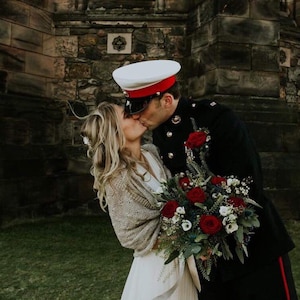 May include: A bride and groom kiss in front of a stone building. The groom is wearing a military uniform with a white hat and red trim. The bride is wearing a white dress and a beige shawl. They are holding a bouquet of red and white flowers.