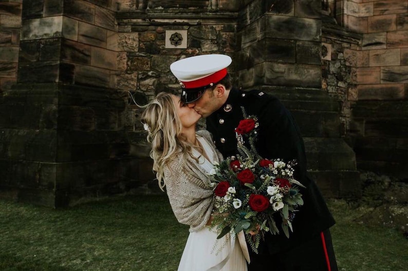 May include: A bride and groom kiss in front of a stone building. The groom is wearing a military uniform with a white hat and red trim. The bride is wearing a white dress and a beige shawl. They are holding a bouquet of red and white flowers.