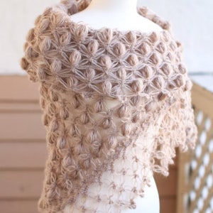May include: A light brown, handmade shawl with a unique, textured design. The shawl is displayed on a mannequin, showcasing its intricate, open-weave pattern and bobble stitch details. The soft, neutral color makes it a versatile accessory.