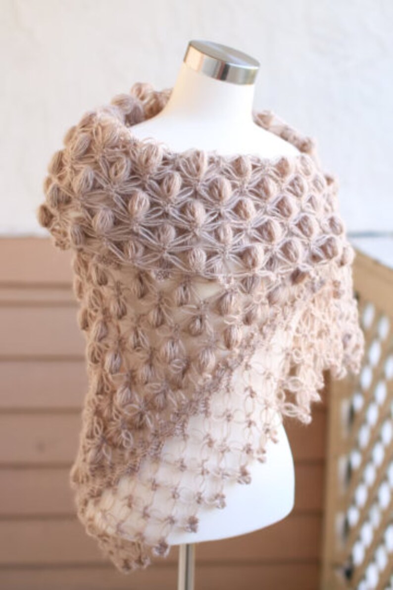 May include: A light brown, handmade shawl with a unique, textured design. The shawl is displayed on a mannequin, showcasing its intricate, open-weave pattern and bobble stitch details. The soft, neutral color makes it a versatile accessory.