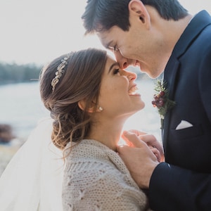 May include: A bride and groom are embracing and looking into each other's eyes. The bride is wearing a white veil and a knitted shawl. The groom is wearing a dark blue suit.