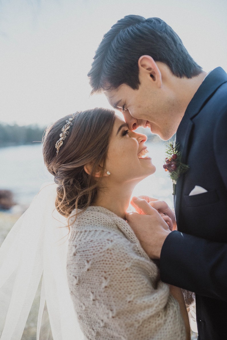 May include: A bride and groom are embracing and looking into each other's eyes. The bride is wearing a white veil and a knitted shawl. The groom is wearing a dark blue suit.
