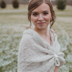 May include: A bride wearing a white knitted shawl with a white satin ribbon tied around the neck. The shawl has a delicate lace pattern. The bride is wearing a white dress and a gold tiara with rhinestones.