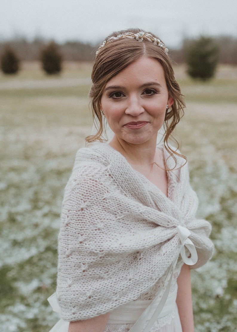 May include: A bride wearing a white knitted shawl with a white satin ribbon tied around the neck. The shawl has a delicate lace pattern. The bride is wearing a white dress and a gold tiara with rhinestones.