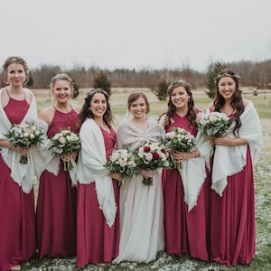 May include: Seven women in burgundy bridesmaid dresses with white shawls stand in a snowy field. They are holding bouquets of white and pink flowers. The woman in the center is wearing a white wedding dress.