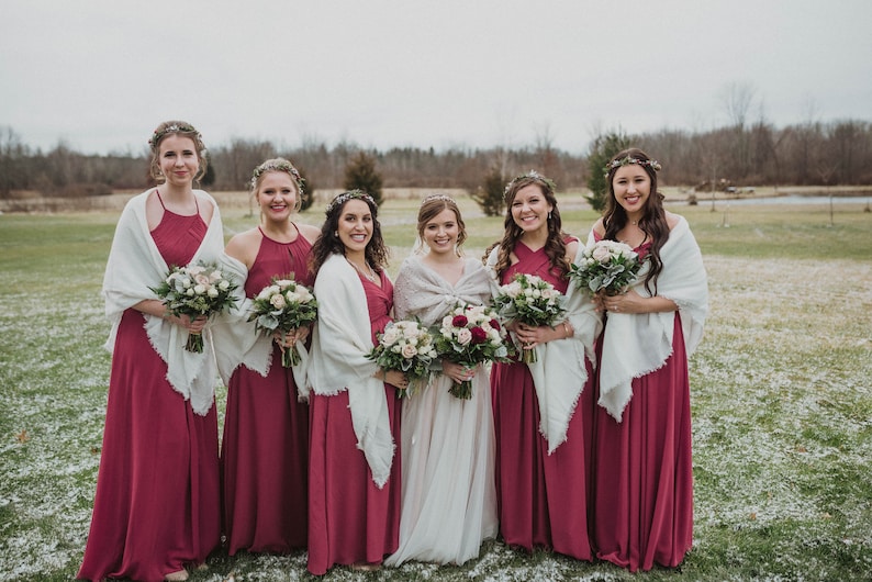 May include: Seven women in burgundy bridesmaid dresses with white shawls stand in a snowy field. They are holding bouquets of white and pink flowers. The woman in the center is wearing a white wedding dress.