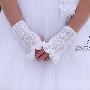 May include: White knitted fingerless gloves with a ruffled edge. The gloves are worn by a person wearing a white dress.