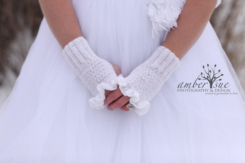 May include: White knitted fingerless gloves with a ruffled edge. The gloves are worn by a person wearing a white dress.