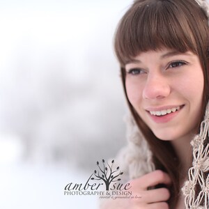 May include: A woman with brown hair and a beige crocheted hood smiles in a snowy outdoor setting. The hood features a floral pattern. The image includes the text "amber sue photography & design rooted & grounded in love."