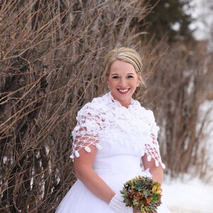 May include: A bride in a white wedding dress with a white crocheted shawl and a bouquet of succulents. The bride is standing in front of a brown bush.