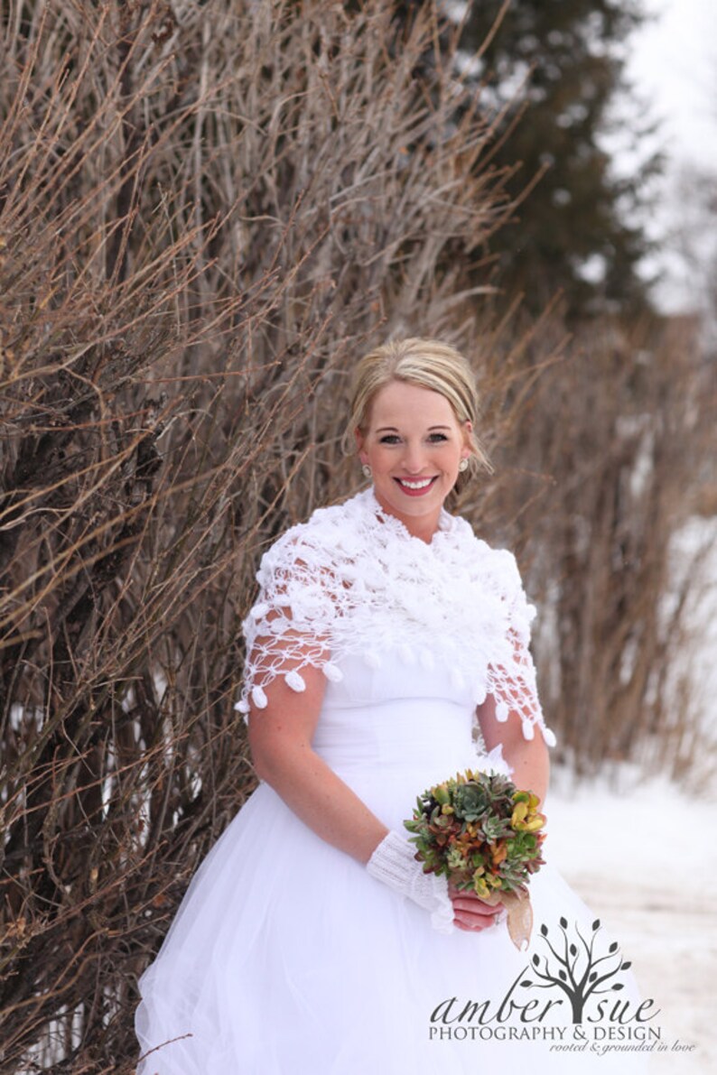 May include: A bride in a white wedding dress with a white crocheted shawl and a bouquet of succulents. The bride is standing in front of a brown bush.