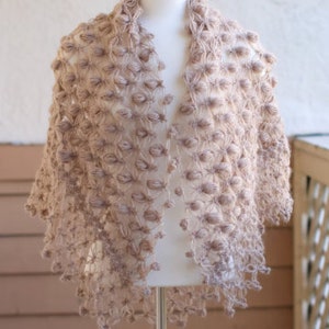 May include: A light brown, open-front shawl with a textured, bobble-like design. The shawl is displayed on a mannequin, showcasing its draped shape and intricate, handmade appearance. The garment is made of a soft, possibly knitted material.