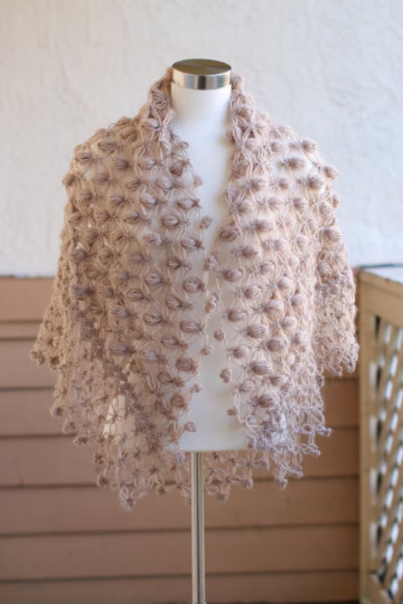 May include: A light brown, open-front shawl with a textured, bobble-like design. The shawl is displayed on a mannequin, showcasing its draped shape and intricate, handmade appearance. The garment is made of a soft, possibly knitted material.