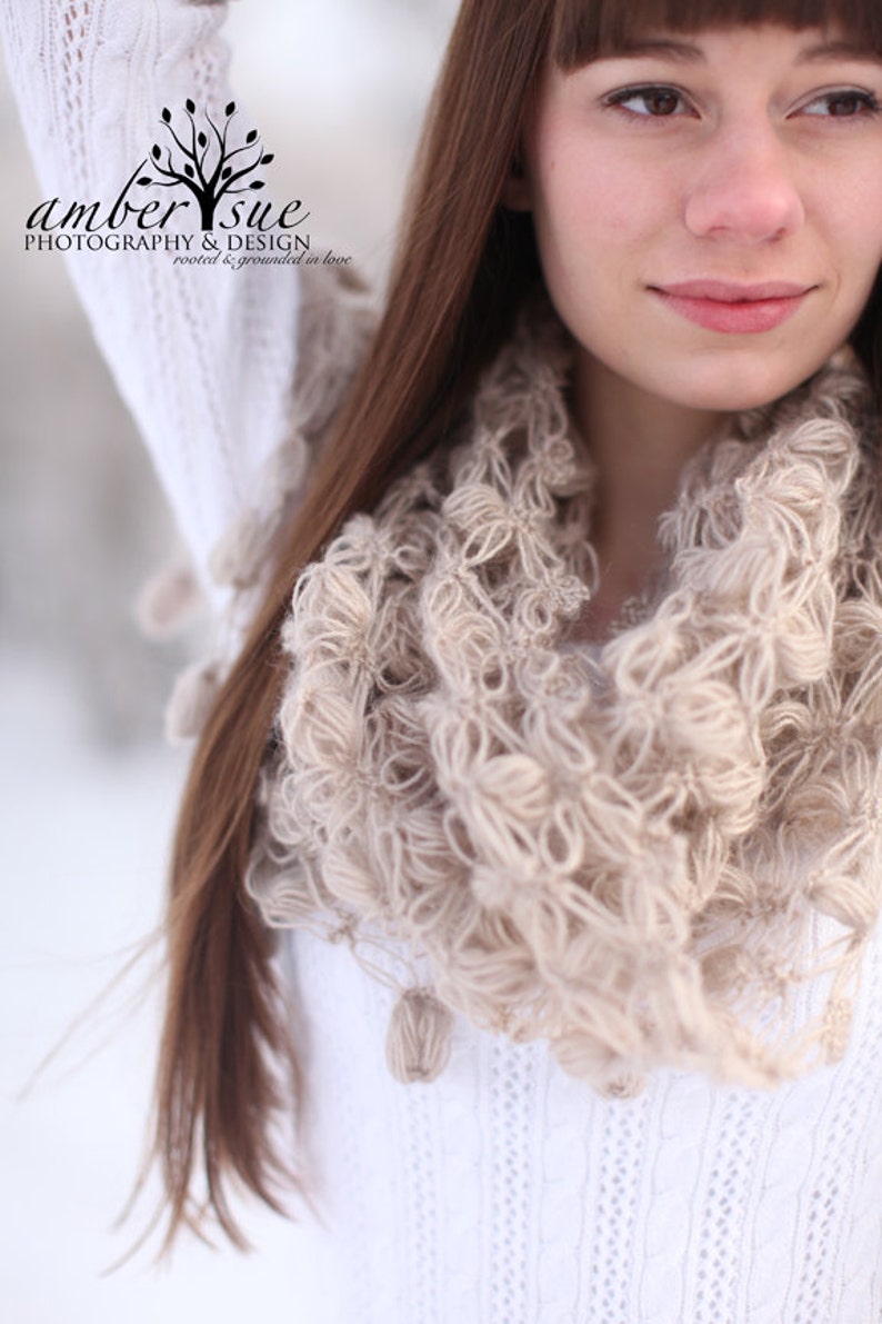 May include: A close-up of a beige infinity scarf with a textured, looped design. The scarf is worn over a white sweater. The image also shows long brown hair and the logo "amber sue photography & design".
