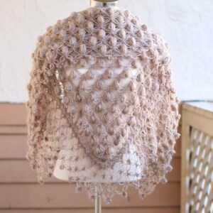 May include: A light brown, handmade shawl with a textured, bobble stitch design. The shawl is displayed on a mannequin and has a triangular shape. The edges are finished with a delicate trim. The overall style is bohemian and cozy.