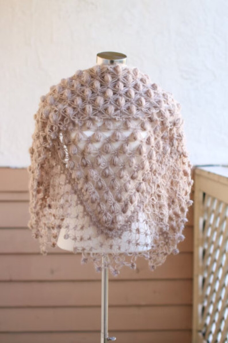 May include: A light brown, handmade shawl with a textured, bobble stitch design. The shawl is displayed on a mannequin and has a triangular shape. The edges are finished with a delicate trim. The overall style is bohemian and cozy.