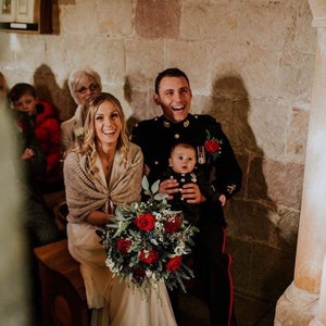 May include: A bride and groom smile at the ceremony, the bride holds a bouquet of red roses and greenery, the groom holds a baby in his arms.