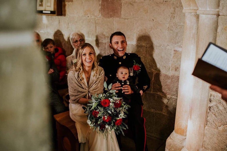 May include: A bride and groom smile at the ceremony, the bride holds a bouquet of red roses and greenery, the groom holds a baby in his arms.
