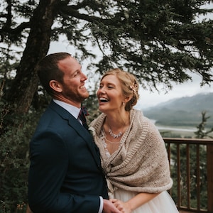 May include: A bride and groom laughing together outdoors. The bride is wearing a white dress with a beige knitted shawl and a silver necklace. The groom is wearing a dark blue suit with a white shirt and a burgundy tie. They are standing in front of a wooden railing with a mountain range and a river in the background.