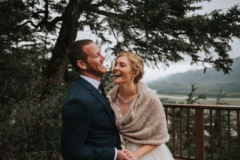 May include: A bride and groom laughing together outdoors. The bride is wearing a white dress with a beige knitted shawl and a silver necklace. The groom is wearing a dark blue suit with a white shirt and a burgundy tie. They are standing in front of a wooden railing with a mountain range and a river in the background.