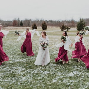 May include: A group of bridesmaids in burgundy dresses and white shawls are laughing and twirling in a snowy field. The bride is in the center wearing a white dress and a white shawl.