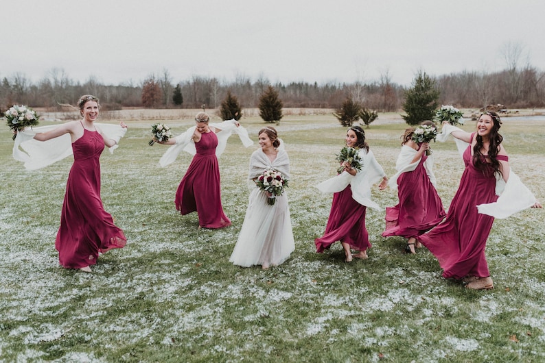 May include: A group of bridesmaids in burgundy dresses and white shawls are laughing and twirling in a snowy field. The bride is in the center wearing a white dress and a white shawl.
