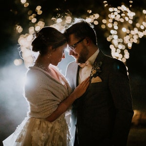 May include: A bride and groom stand close together, looking at each other. The bride is wearing a white dress with a cream-colored shawl. The groom is wearing a dark suit with a light-colored bow tie. They are standing in front of a background of blurred lights.