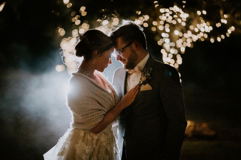 May include: A bride and groom stand close together, looking at each other. The bride is wearing a white dress with a cream-colored shawl. The groom is wearing a dark suit with a light-colored bow tie. They are standing in front of a background of blurred lights.
