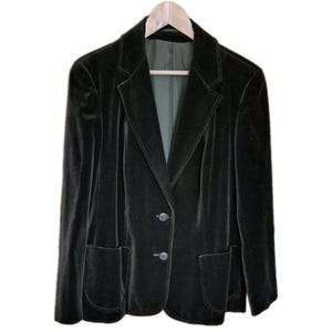 May include: A dark green velvet blazer with a two-button fastening and two front pockets. The jacket is suspended on a wooden hanger. The blazer has a classic lapel design and a tailored fit, against a white backdrop.