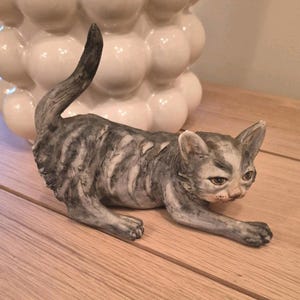 May include: A ceramic cat ornament in a relaxed pose, with grey and white striped fur. The cat has detailed facial features and is positioned on a wooden surface. A white, bubble-shaped decorative object is in the background.