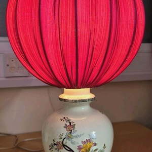 May include: A table lamp featuring a large, spherical, pleated red fabric shade. The base is a white ceramic vase with a floral pattern in pink, yellow, and blue, accented by a decorative green and black geometric border at the top and bottom.
